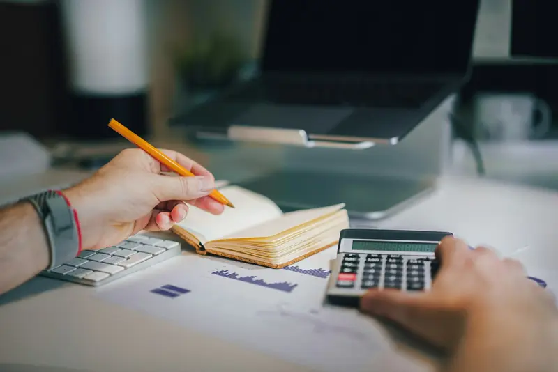 A hand holding a pencil over an open notebook beside a second hand pressing keys on a calculator, with a bar-graph printout and a laptop on the desk.