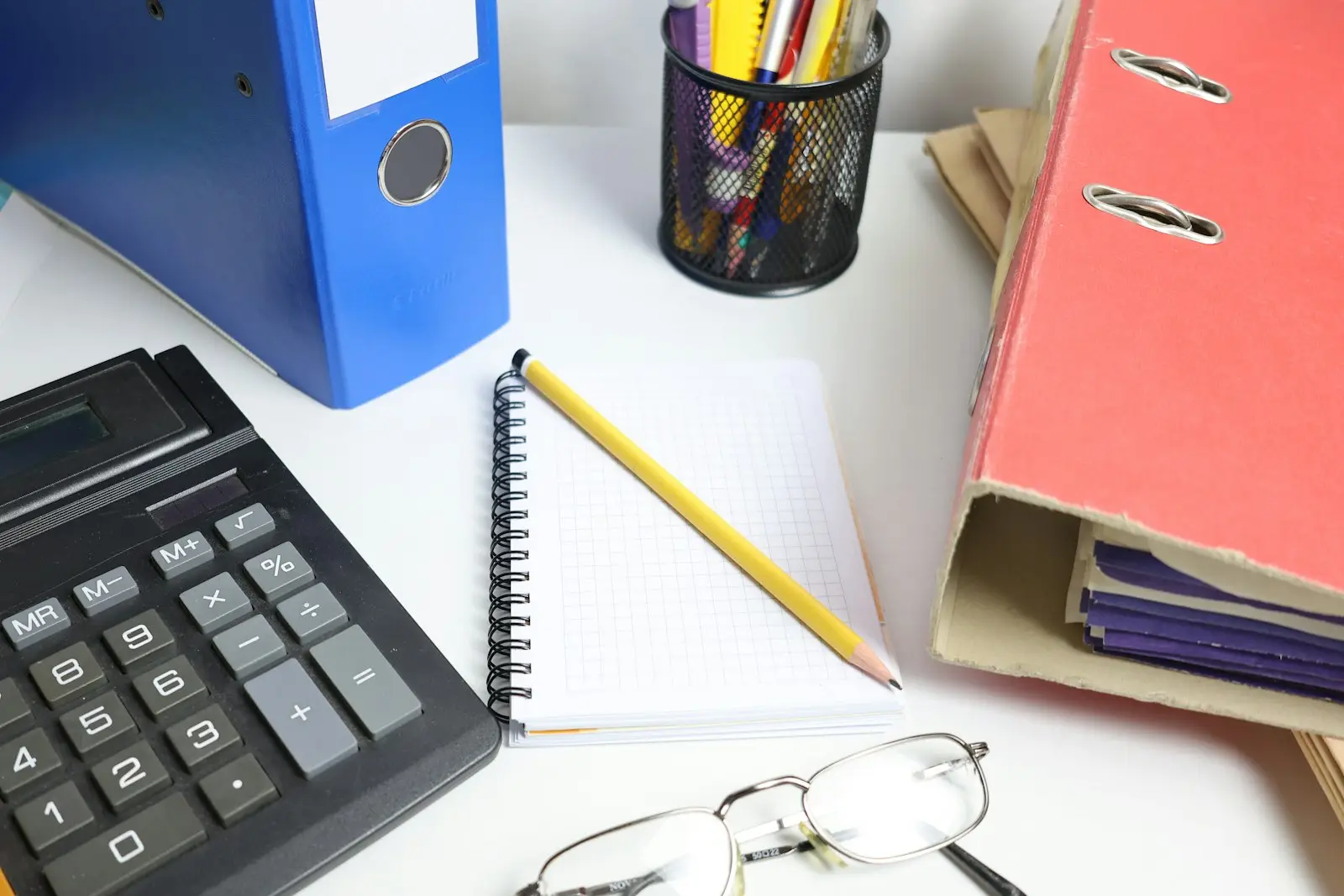 An office desk holding a black calculator, a spiral-bound notebook and pencil, a pen pot, reading glasses and lever-arch folders stuffed with paperwork.