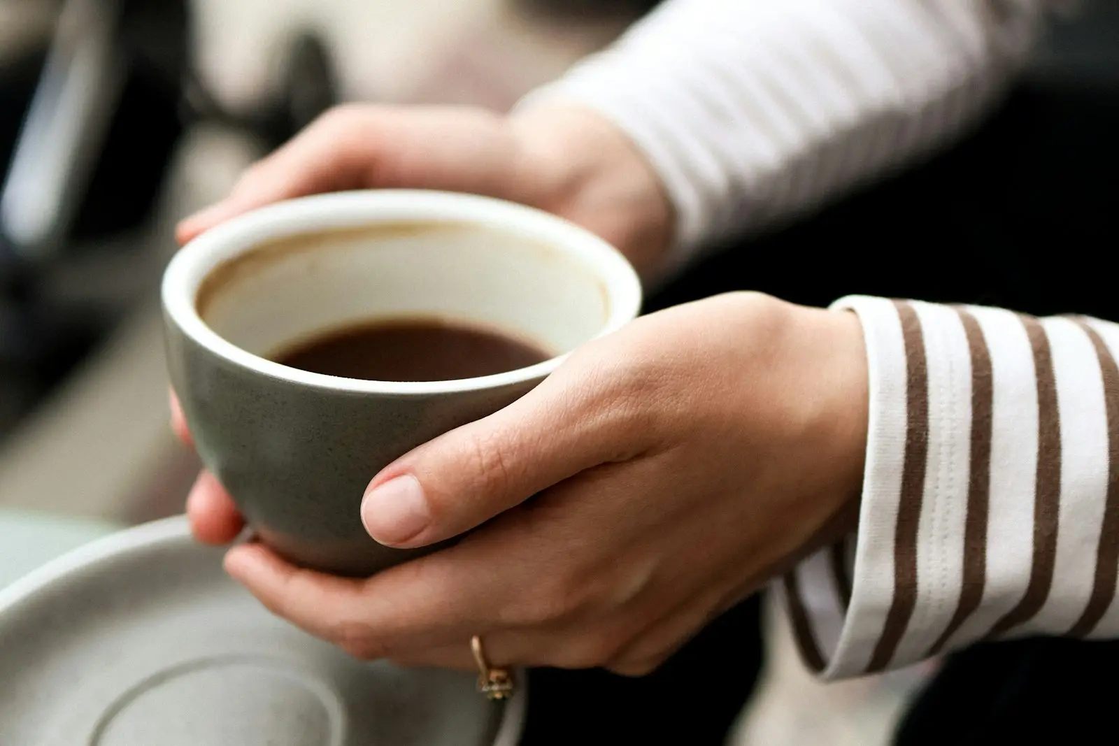 Two hands cupping a warm grey ceramic coffee cup and saucer at a cafe table.