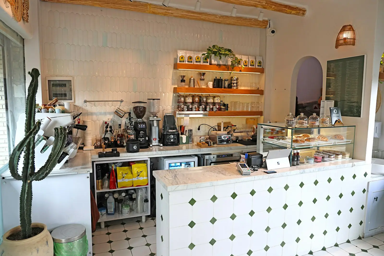 A bright tiled cafe counter with an espresso machine, coffee grinders and open timber shelving stocked with coffee bags and glassware, croissants and pastries under glass domes in the display case.
