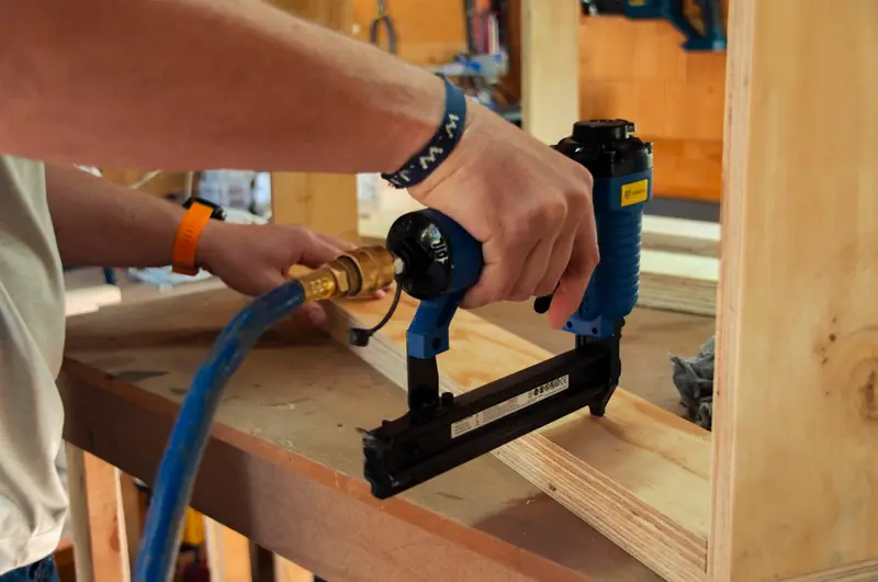 A hand using an air-powered finish nailer to fix plywood panels to a timber-framed cabinet.