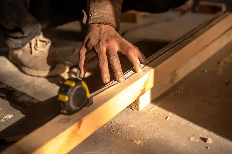 A dusty hand measuring a length of timber framing with a tape measure on a sunlit concrete slab.
