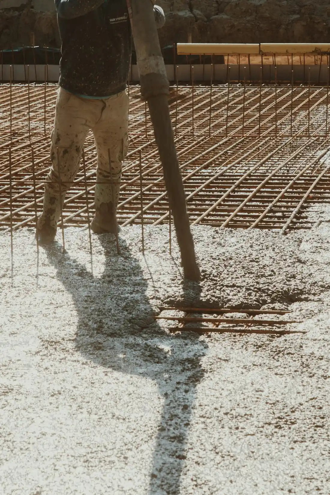 Backlit silhouette of a worker standing on steel reinforcing mesh over a wet concrete pour, holding a vibrating screed pole.