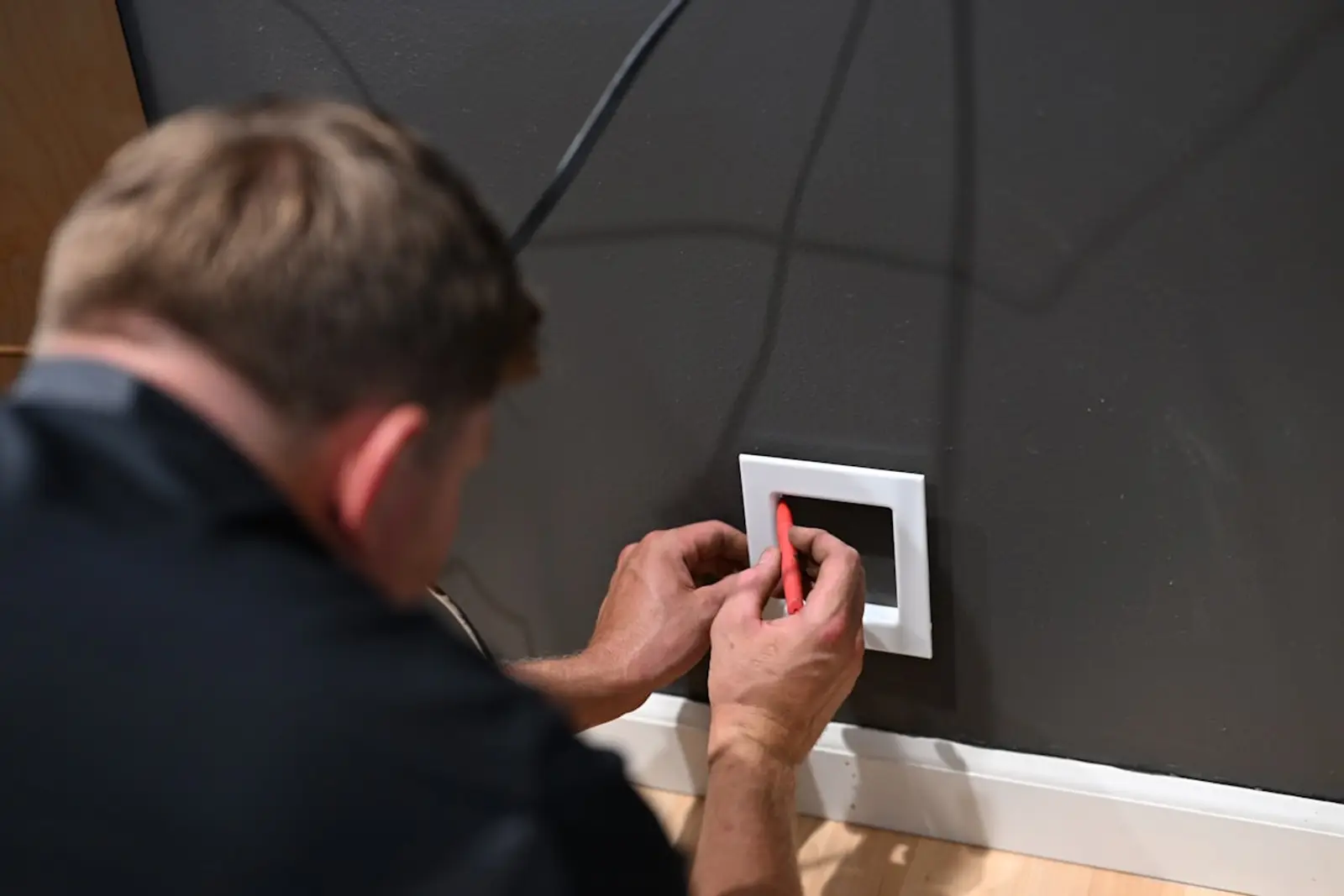 Back of a tradesperson's head and shoulders as they fit a wall power-point plate with a small screwdriver, timber skirting visible below.