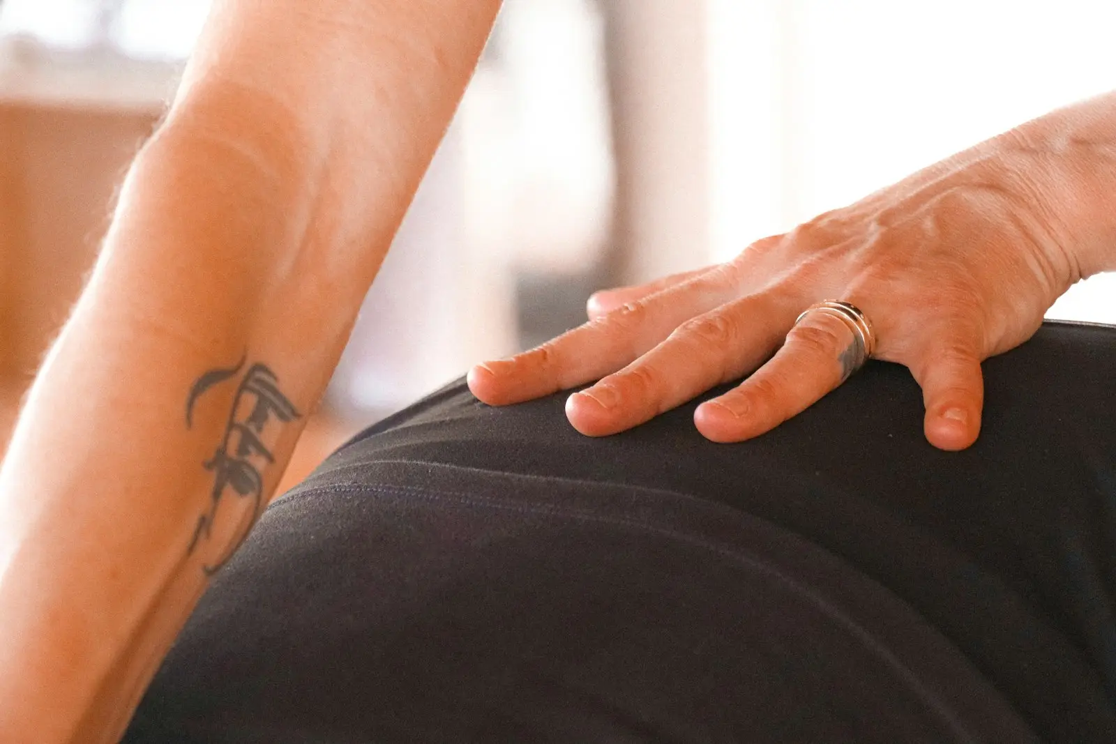 Close-up of a therapist's hand pressing on a client's lower back over dark athletic wear, the client's tattooed forearm braced in the foreground, in a sunlit treatment room.