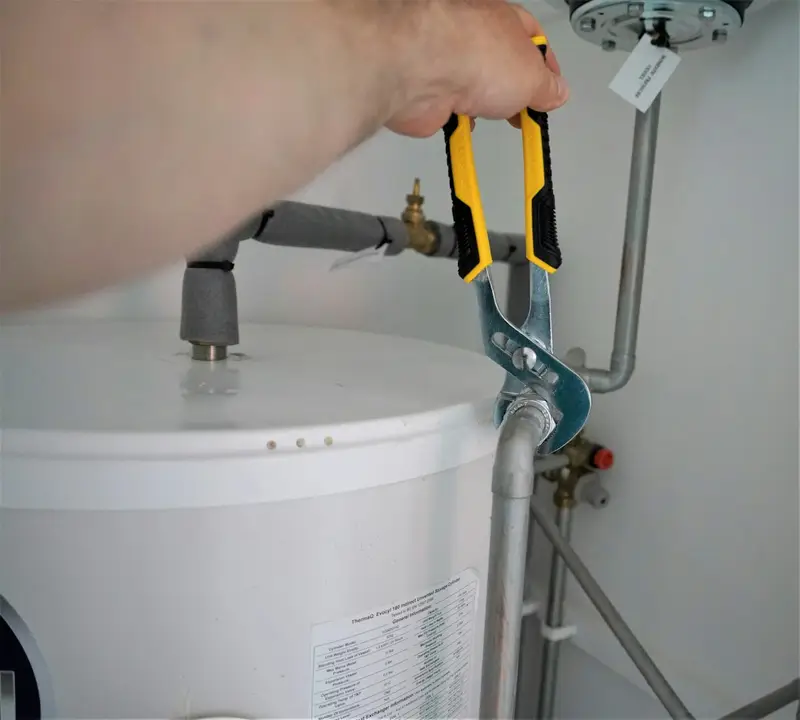 Close-up of yellow-handled pliers tightening a pipe fitting on top of a household hot-water storage cylinder.