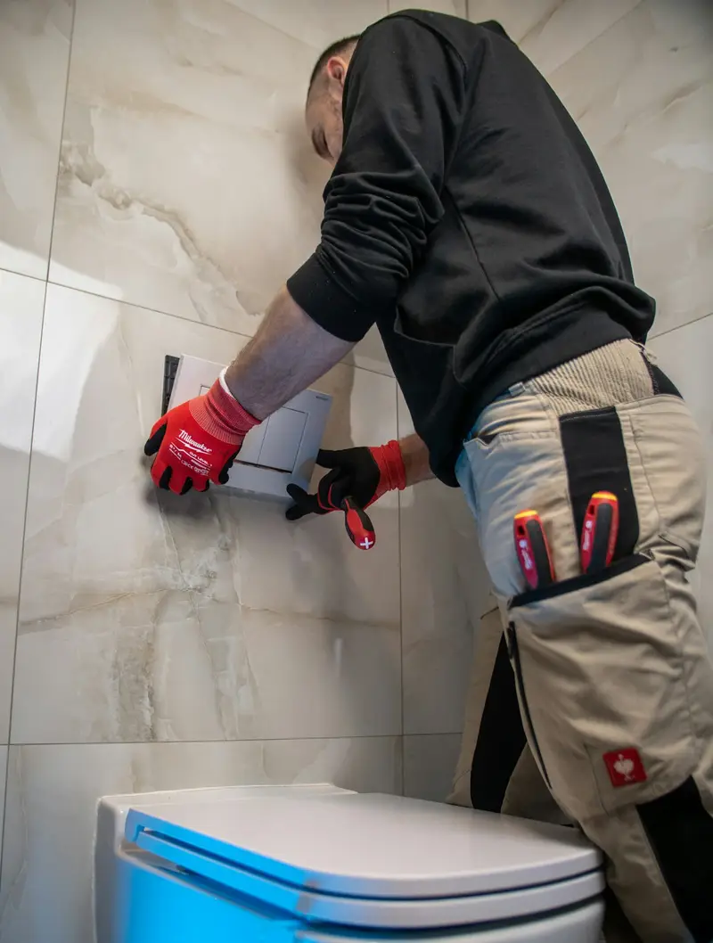 Plumber in red gloves fitting an in-wall toilet cistern flush plate with a screwdriver in a tiled bathroom.