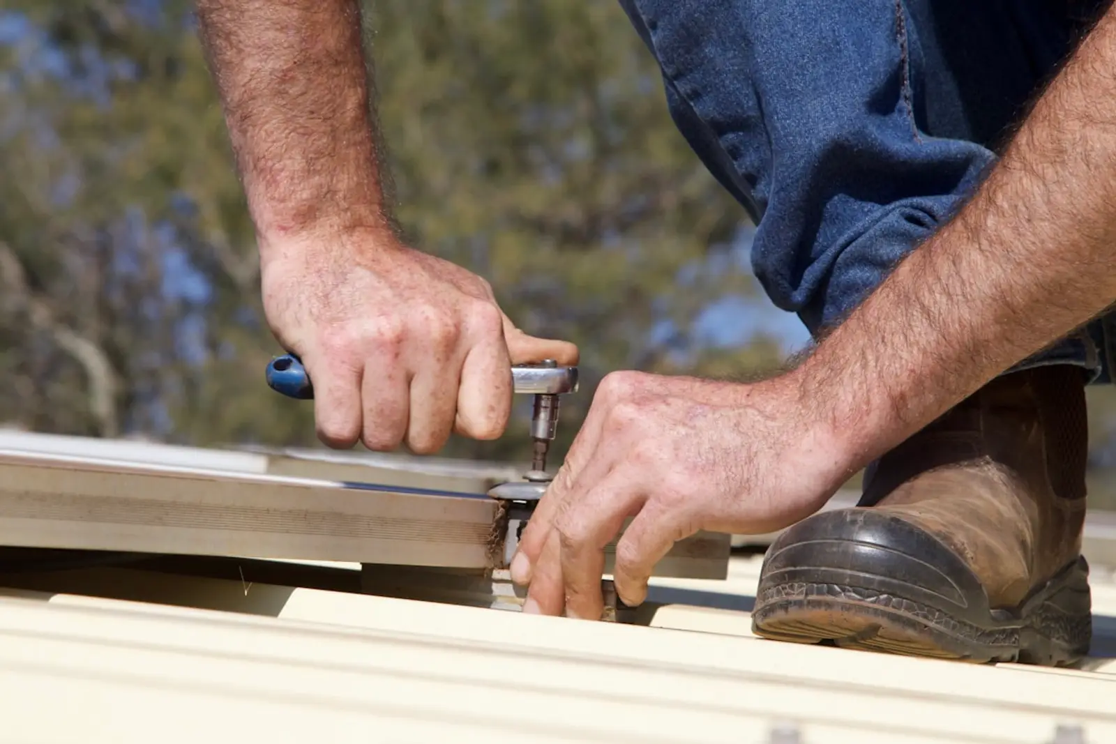 Two hands using a hand crimping tool to join a length of metal roof flashing, trees blurred behind against daylight.